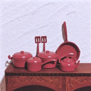 May include: A miniature red kitchen set, including pots, pans, a spatula, and a kettle. The set is arranged on a small, dark wooden cabinet. The items are all a matching shade of red, creating a cohesive and charming aesthetic.