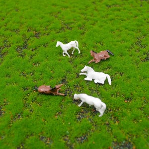 Puede incluir: Figuras de caballos de plástico en miniatura en blanco y marrón están dispersas sobre un lecho de césped artificial verde. Algunos caballos están de pie, mientras que otros están tumbados. La escena evoca un tema pastoral y lúdico.