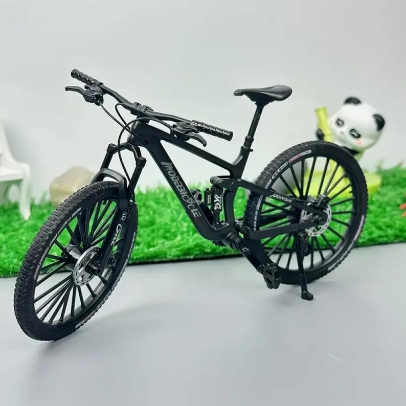 Op de afbeelding: Een zwarte miniatuur mountainbike met de tekst "MODEBIKE CYCLE" op het frame. De fiets heeft zwarte banden, een zwart zadel en een zwart stuur. De fiets staat op een groene ondergrond.