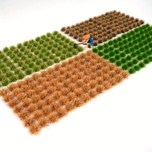 Puede incluir: Modelo en miniatura de un campo dividido en cuatro secciones de diferentes colores: verde claro, verde oscuro, marrón y beige. Una pequeña figura con ropa azul y naranja se muestra trabajando en el campo con una herramienta. La escena está ambientada sobre un fondo blanco.