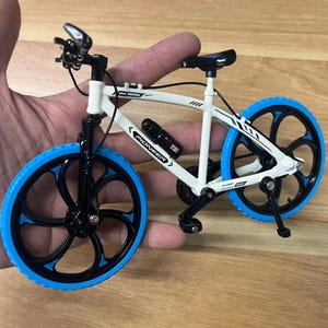 Op de afbeelding: Een miniatuur witte fiets met zwarte en blauwe accenten. De fiets heeft blauwe banden, zwarte velgen en een zwarte waterfles met het woord "PIONEER". De fiets wordt in de hand van een persoon gehouden.