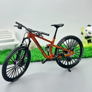 Könnte beinhalten: Ein orangefarbenes und schwarzes Miniatur-Mountainbike mit dem Text "MODEBICYCLE" auf dem Rahmen. Das Fahrrad hat schwarze Reifen, einen schwarzen Sattel und einen schwarzen Lenker. Das Fahrrad steht auf einem Ständer.