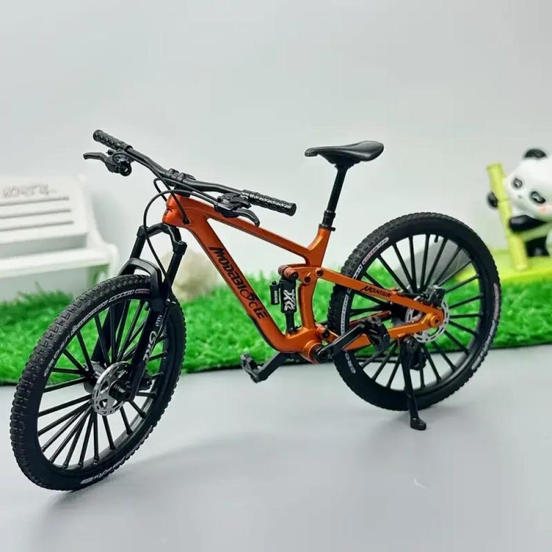 Op de afbeelding: Een oranje en zwarte miniatuur mountainbike met de tekst "MODEBICYCLE" op het frame. De fiets heeft zwarte banden, een zwart zadel en een zwart stuur. De fiets staat op een standaard.