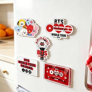 Pode incluir: Uma coleção de ímanes coloridos com personagens de desenhos animados e o texto "BTS World Tour Arirang 2026" em vermelho e branco. Os ímanes estão fixados na porta branca de um frigorífico, com um pano de pratos xadrez vermelho e branco.