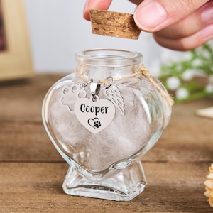 Pode incluir: Um frasco de vidro transparente em forma de coração com uma rolha de cortiça. O frasco contém penugem branca e um pingente de prata com o nome "Cooper" e uma pegada. Uma pegada e um pingente de asa de anjo também estão anexados.