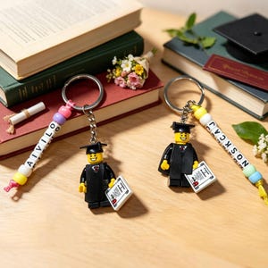 Könnte beinhalten: Zwei Schlüsselanhänger zum Thema Abschlussfeier mit Lego-Figuren in Abschlusskleidung und -kappen. Ein Schlüsselanhänger schreibt "AIVILO" und der andere "NOSKCAJ" mit bunten Perlen. Die Schlüsselanhänger sind in der Nähe von Büchern und einer Abschlusskappe platziert.