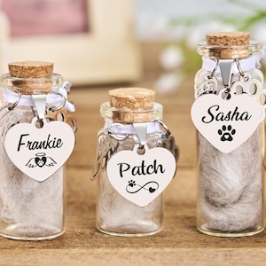 Könnte beinhalten: Drei transparente Gedenkgläser mit Korken, die jeweils das Fell eines Haustiers enthalten. Jedes Glas hat ein herzförmiges Etikett mit dem Namen des Haustiers: Frankie, Patch und Sasha. Das Frankie-Etikett hat Engelsflügel, Patch hat einen Pfotenabdruck mit Unendlichkeitszeichen.