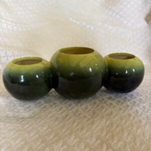 May include: Three round, ceramic candle holders in shades of green, arranged in a connected cluster. The holders have a glossy finish and a gradient effect, transitioning from a lighter yellow-green at the top to a darker, almost black-green at the base. The background is a white, textured surface.