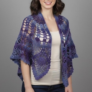 May include: A handmade, open-weave shawl in shades of blue and purple. The shawl is crocheted with a lacy pattern and has a draped, open front design. The garment is worn over a white top and blue jeans.