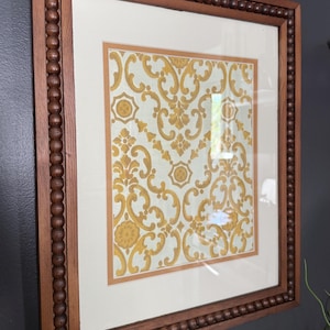 Puede incluir: Obra de arte enmarcada con un fondo crema y un patrón floral dorado repetido. La obra está encerrada en un marco de madera marrón oscuro con un borde de cuentas. El marco es rectangular y tiene un passe-partout crema.