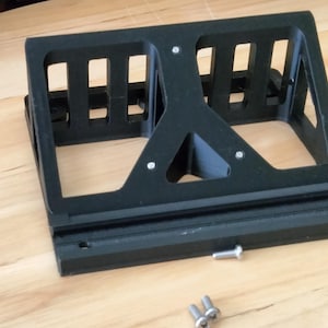 May include: Black, rectangular, 3D-printed object with a series of rectangular cutouts. The object has a triangular support in the center and a flat base. Several screws are visible near the base. The object appears to be a bracket or holder.