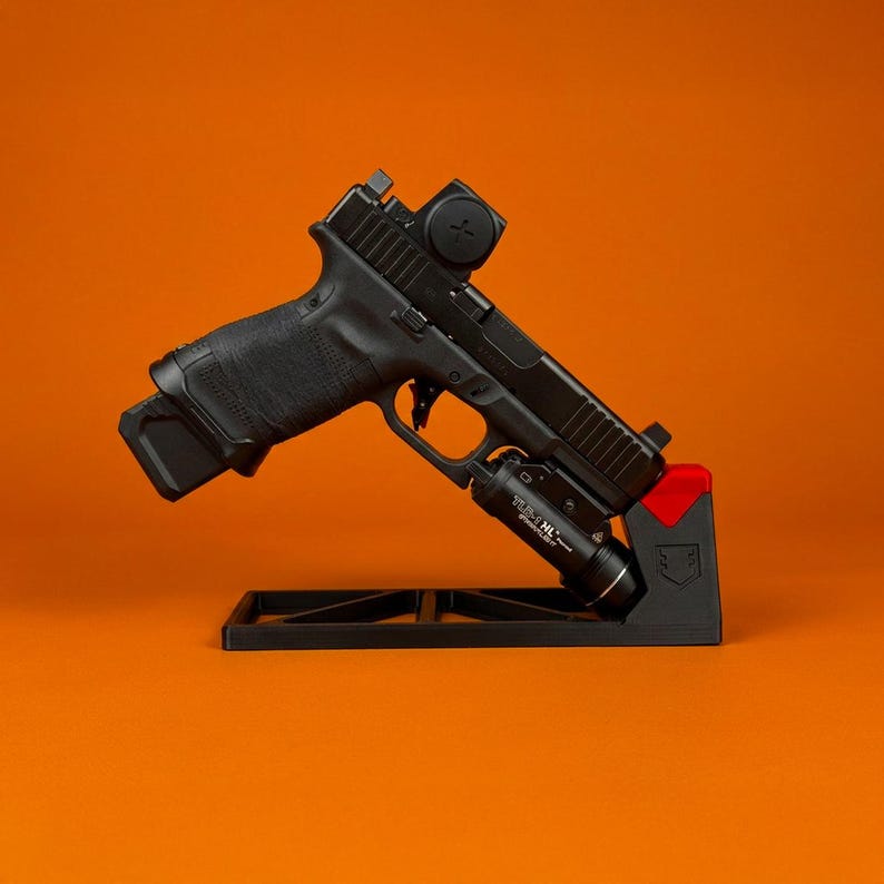 Pode incluir: Uma pistola semiautom&aacute;tica preta montada num suporte de exposi&ccedil;&atilde;o preto e vermelho. A arma possui uma mira de ponto vermelho na corredi&ccedil;a e uma lanterna t&aacute;tica Streamlight TLR-1 HL fixada no trilho de acess&oacute;rios sob o cano.