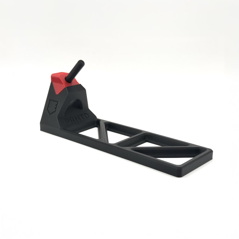 Pode incluir: Um suporte de pl&aacute;stico preto com uma base retangular apresentando um padr&atilde;o de recortes triangulares. O suporte tem uma pe&ccedil;a de destaque vermelha no topo e um pino cil&iacute;ndrico preto. A palavra PRNTD est&aacute; gravada na lateral do suporte.