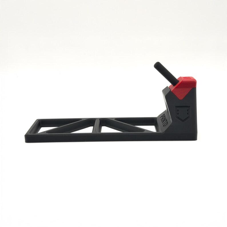 Pode incluir: Um suporte de pl&aacute;stico preto com uma base retangular apresentando um padr&atilde;o geom&eacute;trico recortado. O suporte possui uma se&ccedil;&atilde;o vertical com uma tampa vermelha e um pino preto inclinado. A palavra PRNTD est&aacute; gravada na lateral do suporte.