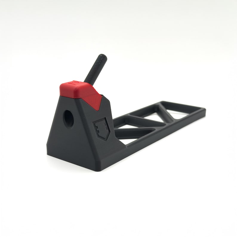 Pode incluir: Um suporte de pl&aacute;stico preto fosco com uma base triangular, um detalhe vermelho no topo e um poste preto inclinado. A estrutura inclui uma abertura circular oca na face frontal e uma se&ccedil;&atilde;o traseira plana e alongada com recortes geom&eacute;tricos.