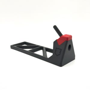 Pode incluir: Um suporte de pl&aacute;stico preto fosco com uma base longa e plana com um padr&atilde;o de recortes triangulares. A se&ccedil;&atilde;o vertical inclui uma pe&ccedil;a de destaque vermelha no topo, um pequeno pino cil&iacute;ndrico preto e um logotipo em forma de escudo.
