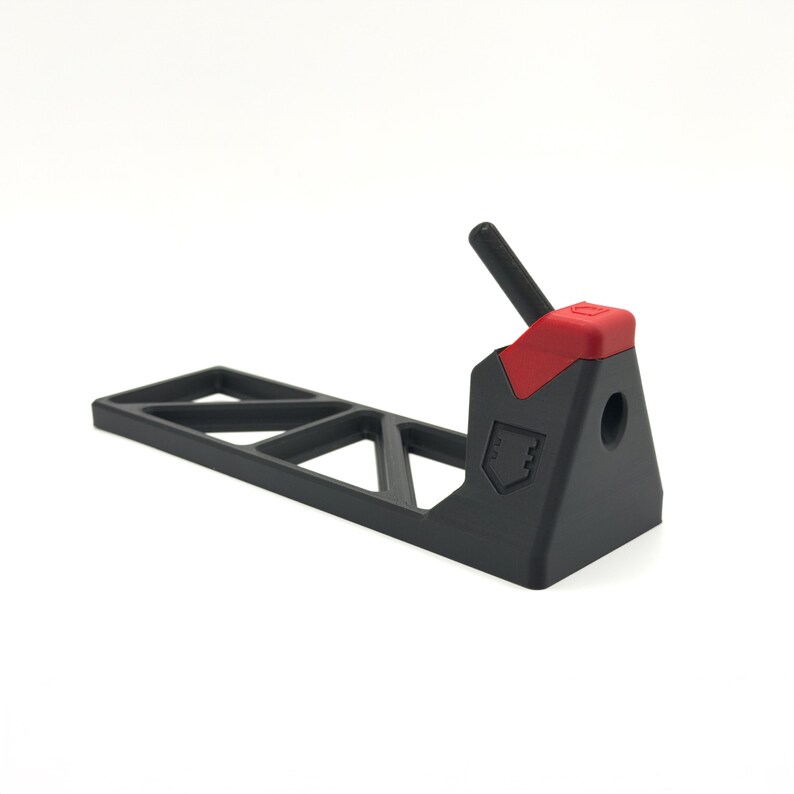 Pode incluir: Um suporte de pl&aacute;stico preto fosco com uma base longa e plana com um padr&atilde;o de recortes triangulares. A se&ccedil;&atilde;o vertical inclui uma pe&ccedil;a de destaque vermelha no topo, um pequeno pino cil&iacute;ndrico preto e um logotipo em forma de escudo.