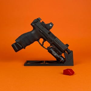 Pode incluir: Uma pistola semiautom&aacute;tica preta montada num suporte de exposi&ccedil;&atilde;o preto. A arma de fogo possui uma mira de ponto vermelho, uma lanterna t&aacute;tica preta acoplada ao trilho de acess&oacute;rios e uma empunhadura texturizada. Um pequeno componente de pl&aacute;stico vermelho encontra-se pr&oacute;ximo.