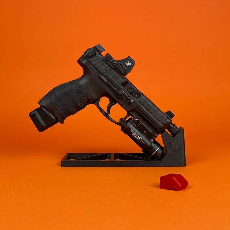 Pode incluir: Uma pistola semiautom&aacute;tica preta montada num suporte de exposi&ccedil;&atilde;o preto. A arma de fogo possui uma mira de ponto vermelho, uma lanterna t&aacute;tica preta acoplada ao trilho de acess&oacute;rios e uma empunhadura texturizada. Um pequeno componente de pl&aacute;stico vermelho encontra-se pr&oacute;ximo.