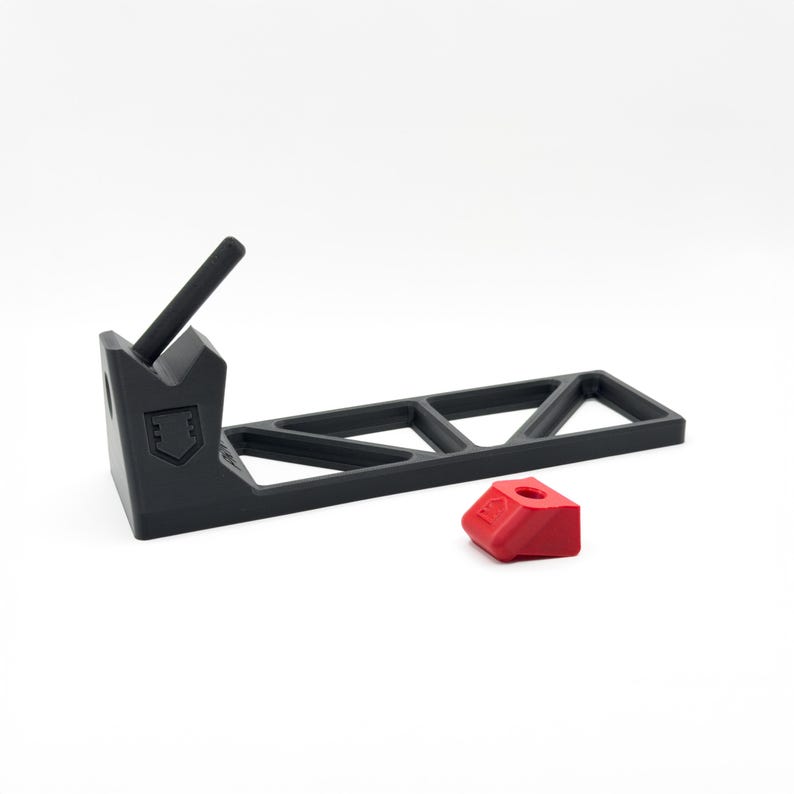 Pode incluir: Um suporte preto em forma de L com uma base de treli&ccedil;a e um bra&ccedil;o de apoio inclinado com um emblema em forma de escudo. Um pequeno componente vermelho separado em forma de cunha, com um orif&iacute;cio central e uma seta em relevo, est&aacute; &agrave; frente.