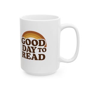 Puede incluir: Taza de cerámica blanca con un asa grande. La taza presenta el texto "GOOD DAY TO READ" en letras marrones con un diseño de arcoíris marrón y amarillo encima del texto. La taza está diseñada para bebidas calientes.
