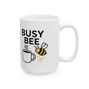 Puede incluir: Taza de cerámica blanca con las palabras "BUSY BEE" en negro, una abeja de dibujos animados y una taza de café humeante. La abeja es amarilla y negra, y la taza de café es negra y blanca. La taza tiene un asa grande.
