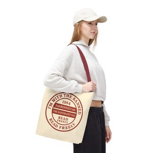 Puede incluir: Bolso tote de color crema con una correa burdeos y un diseño de texto circular. El texto dice "I'M WITH THE BANNED" en el borde, con "1984", "FAHRENHEIT 451" y "THE HANDMAID'S TALE" en el centro. Debajo, dice "READ FREELY".