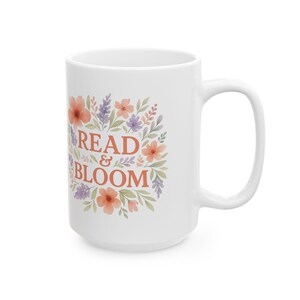 Puede incluir: Taza de cerámica blanca con las palabras "READ & BLOOM" en naranja, rodeada de un diseño floral en tonos naranja, morado y verde. La taza tiene un asa grande.
