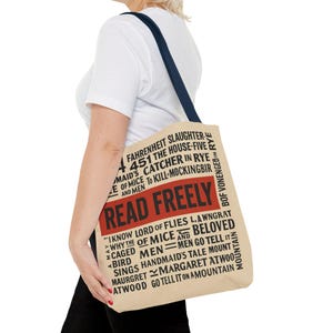 Puede incluir: Bolso tote beige con una correa azul marino, con el texto "READ FREELY" en rojo. La bolsa enumera varios títulos de libros en negro, incluyendo "Fahrenheit 451" y "Catcher in the Rye".
