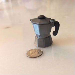 Peut inclure: Une petite cafetière italienne grise avec une poignée et un dessus noirs, à côté d'une pièce de monnaie. La cafetière miniature a un design classique avec une base hexagonale et un corps strié.
