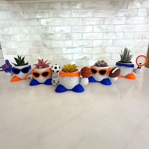 May include: A collection of whimsical, sports-themed succulent planters. Each planter is white with blue and orange accents, sunglasses, and holds a different sport-related item, including a foam finger, soccer ball, basketball, and tennis racket.