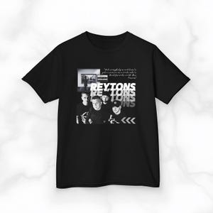 May include: Black t-shirt featuring a grayscale image of a band with the name "REYTONS" in a layered, white font. The shirt also includes a quote and directional arrows.