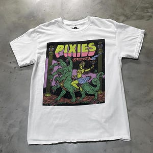 Pode incluir: T-shirt branca com um gráfico colorido de uma criatura mítica e uma mulher. O gráfico inclui a palavra "PIXIES" em verde e rosa, com "US TOUR 2023" abaixo. A t-shirt é feita de um material macio e confortável.