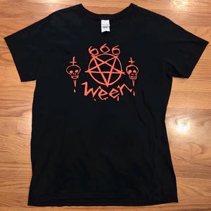 Puede incluir: Una camiseta negra de manga corta con un estampado gráfico de color naranja brillante en el pecho. El diseño incluye un pentagrama con el número 666 encima, flanqueado por dos calaveras con cruces y la palabra WEEN en una fuente estilizada.