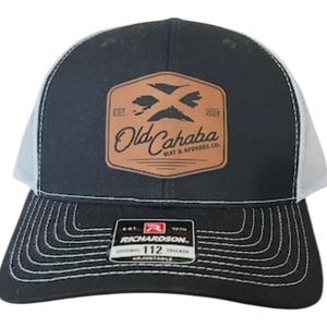 May include: Black and white trucker cap with a curved peak and mesh back. The front has a brown leather patch with the text "Old Cahaba HAT & APPAREL CO." and a graphic of two crossed birds. The cap is adjustable.