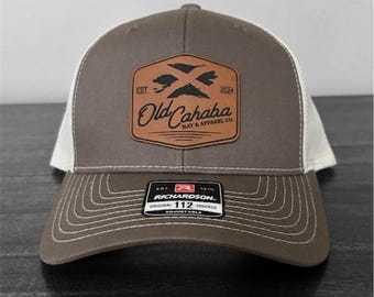 Könnte beinhalten: Braune und weiße Trucker-Cap mit einem Lederaufnäher mit dem Text "Old Cahaba Hat & Apparel Co." und einem gekreuzten Pfeildesign. Die Kappe hat einen gebogenen Schirm und einen verstellbaren Druckknopfverschluss.