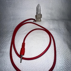 Può includere: Un ciondolo di cristallo di quarzo trasparente avvolto con filo color argento, collegato a un cavo audio rosso con un jack rosso da 3,5 mm. Il cristallo è alto circa 5 cm. Il cavo è arrotolato su una superficie bianca strutturata.