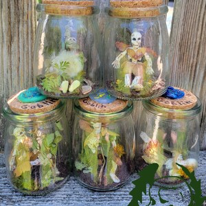 May include: Five glass jars with cork lids, each containing a miniature fantasy scene. The jars feature small figurines, plants, and decorative elements. The lids are adorned with colorful embellishments. The word "Goblin" is visible on some of the lids.