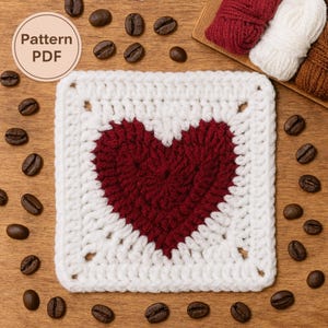 Peut inclure: Un dessous de verre carré crocheté avec un motif de cœur rouge au centre, entouré d'une bordure blanche. Les mots "Pattern PDF" sont dans un cercle en haut à gauche. Des grains de café sont dispersés autour du dessous de verre sur une surface en bois.