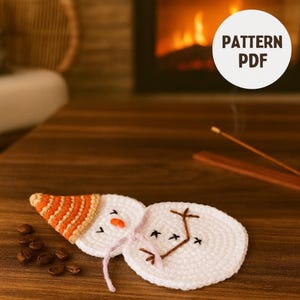 Peut inclure: Sous-verres faits à la main en forme de bonhomme de neige au crochet. Le bonhomme de neige a un corps blanc, un chapeau orange et blanc et des détails brodés. Les dessous de verre sont sur une surface en bois avec des grains de café. Le texte "PATTERN PDF" est visible.