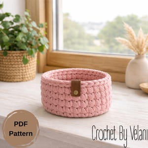 May include: A round, light pink crochet basket with a brown leather strap and button closure. The basket is sitting on a white surface. The text "PDF Pattern" and "Crochet By Velani" are visible.