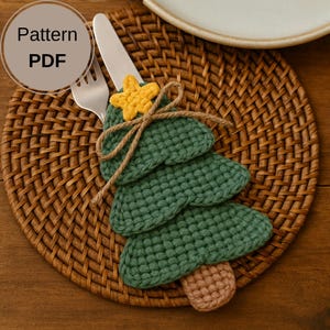 Könnte beinhalten: Ein grüner gehäkelter Weihnachtsbaum-Besteckhalter mit einem gelben Stern und einer braunen Schnurschleife. Der Halter befindet sich auf einem gewebten Platzset mit Gabel und Messer. Der Text "Pattern PDF" befindet sich in einem Kreis.