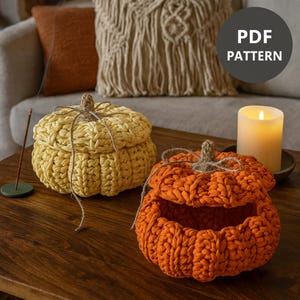 May include: Two handmade, crochet pumpkin-shaped storage containers. One is open, revealing the interior, and the other is closed. The pumpkins are made of thick yarn in yellow and orange. A candle and the text "PDF PATTERN" are also visible.
