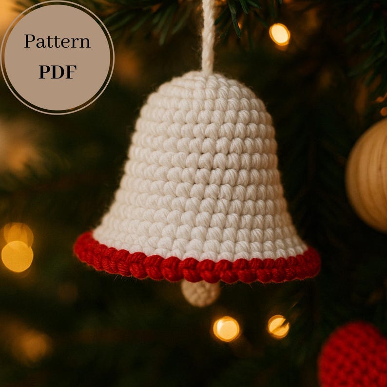 Op de afbeelding: Een gehaakte bel-ornament met een witte body en een rode rand. De bel hangt aan een witte draad tegen een wazige achtergrond van een kerstboom. De tekst "Pattern PDF" staat in een cirkel linksboven.