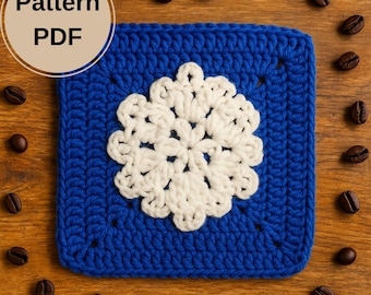 Op de afbeelding: Een vierkant, blauw gehaakt stuk met een wit bloemmotief in het midden. De tekst "Pattern PDF" staat in een cirkel. Koffiebonen zijn verspreid rond het gehaakte vierkant op een houten oppervlak.