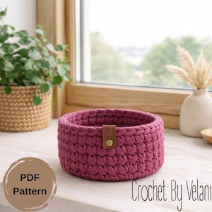 May include: A round, crocheted basket in a deep rose color, featuring a brown leather strap with a gold button closure. The basket is handmade and sits on a white surface. The text "PDF Pattern" is visible on a brown circle.