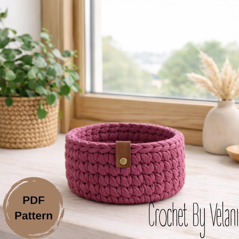 May include: A round, crocheted basket in a deep rose color, featuring a brown leather strap with a gold button closure. The basket is handmade and sits on a white surface. The text "PDF Pattern" is visible on a brown circle.