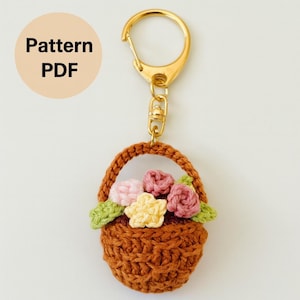 May include: A crochet keychain with a small brown basket filled with pink, yellow, and green flowers. The keychain has a gold-coloured clasp and ring. The image also includes the text "Pattern PDF".