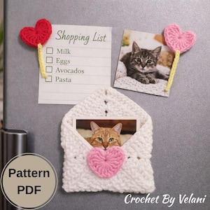 May include: A white crocheted envelope with a pink heart and a photo of a cat. A shopping list and two photos of cats are attached with crocheted heart magnets. The text "Pattern PDF" and "Crochet By Velani" are visible.