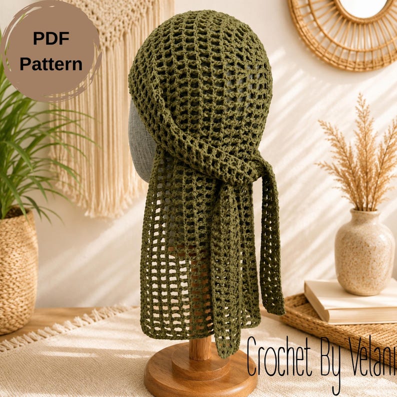 May include: An olive green crochet headscarf featuring an open mesh stitch pattern with two long ties hanging down the back. The head covering is displayed on a mannequin head stand. The image includes a circular badge with the text PDF Pattern.